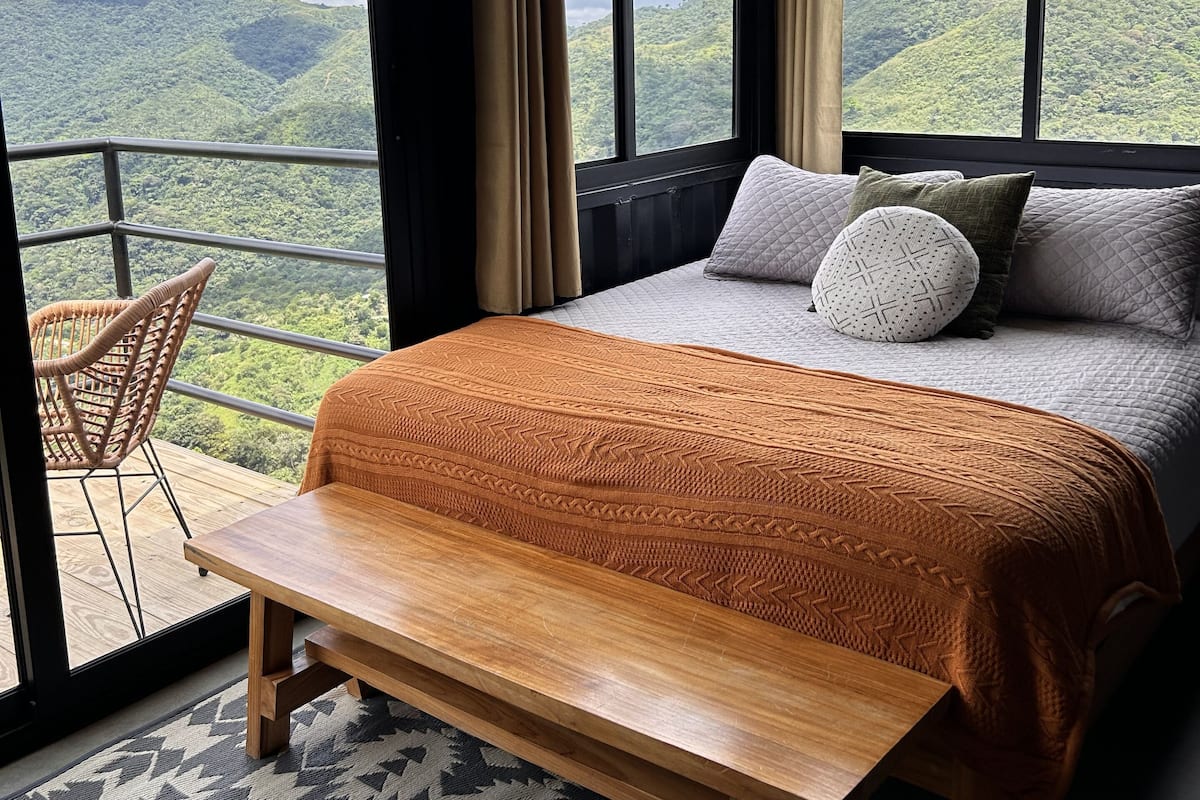 Une chambre avec un grand lit, un banc en bois et une vue sur un paysage montagneux, visible à travers de grandes fenêtres.