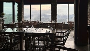 A dining area with a large table and wicker chairs, large windows with a mountain view, and stone pillars. - Nomada Republic Hotel El Valle  (El Valle de Anton)