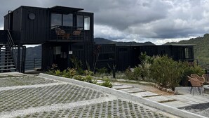 A modern black building with a glass balcony, surrounded by a garden with plants and a paved area with chairs. - Nomada Republic Hotel El Valle  (El Valle de Anton)