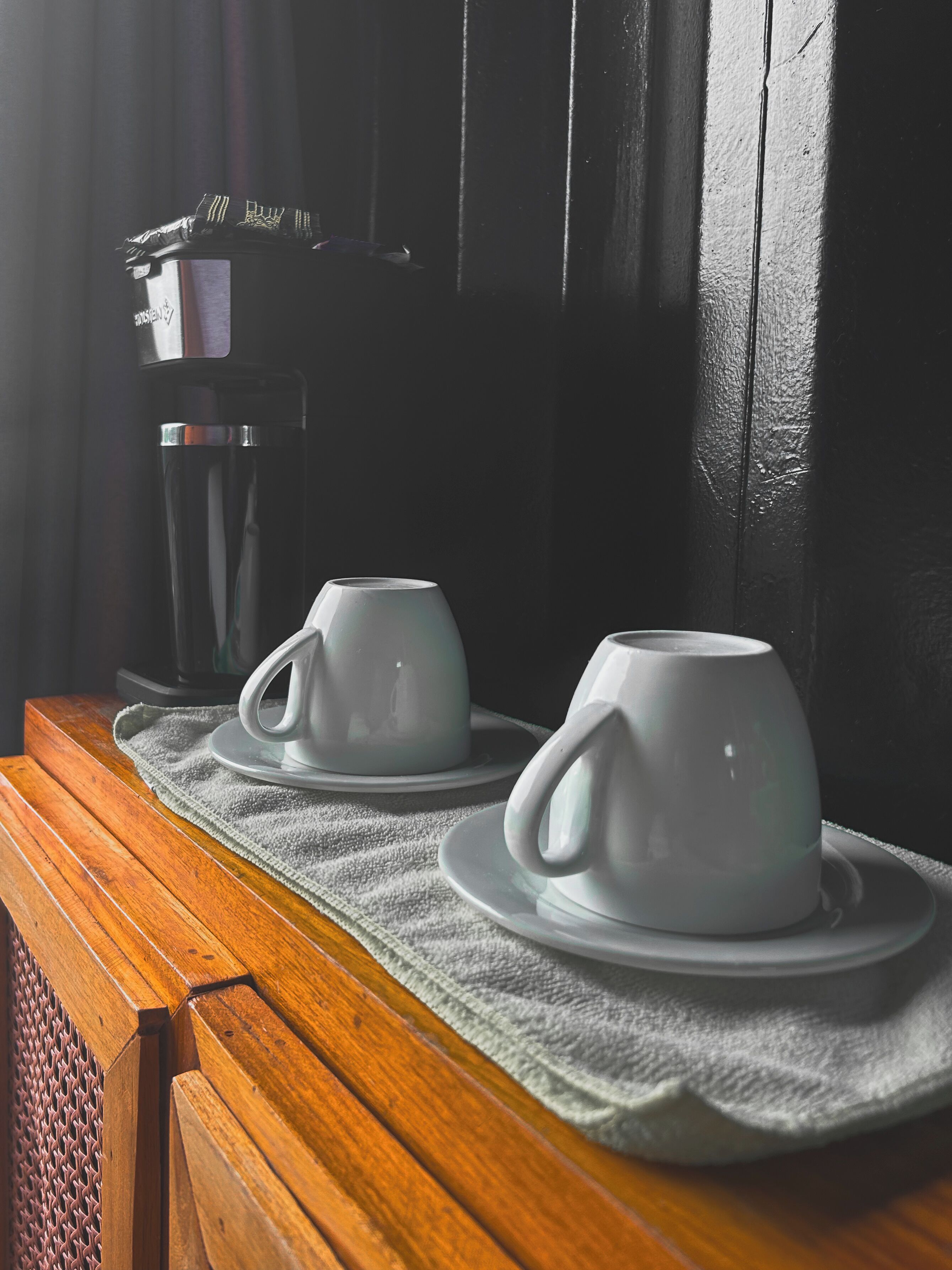 Dos tazas blancas sobre una mesa de madera con un mantel, junto a una cafetera.