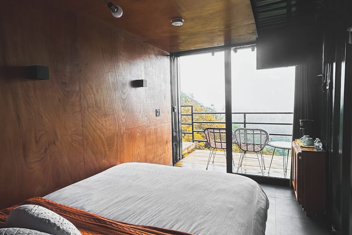 Une chambre avec un grand lit, des murs en bois, un balcon avec une chaise et une vue sur l’extérieur.