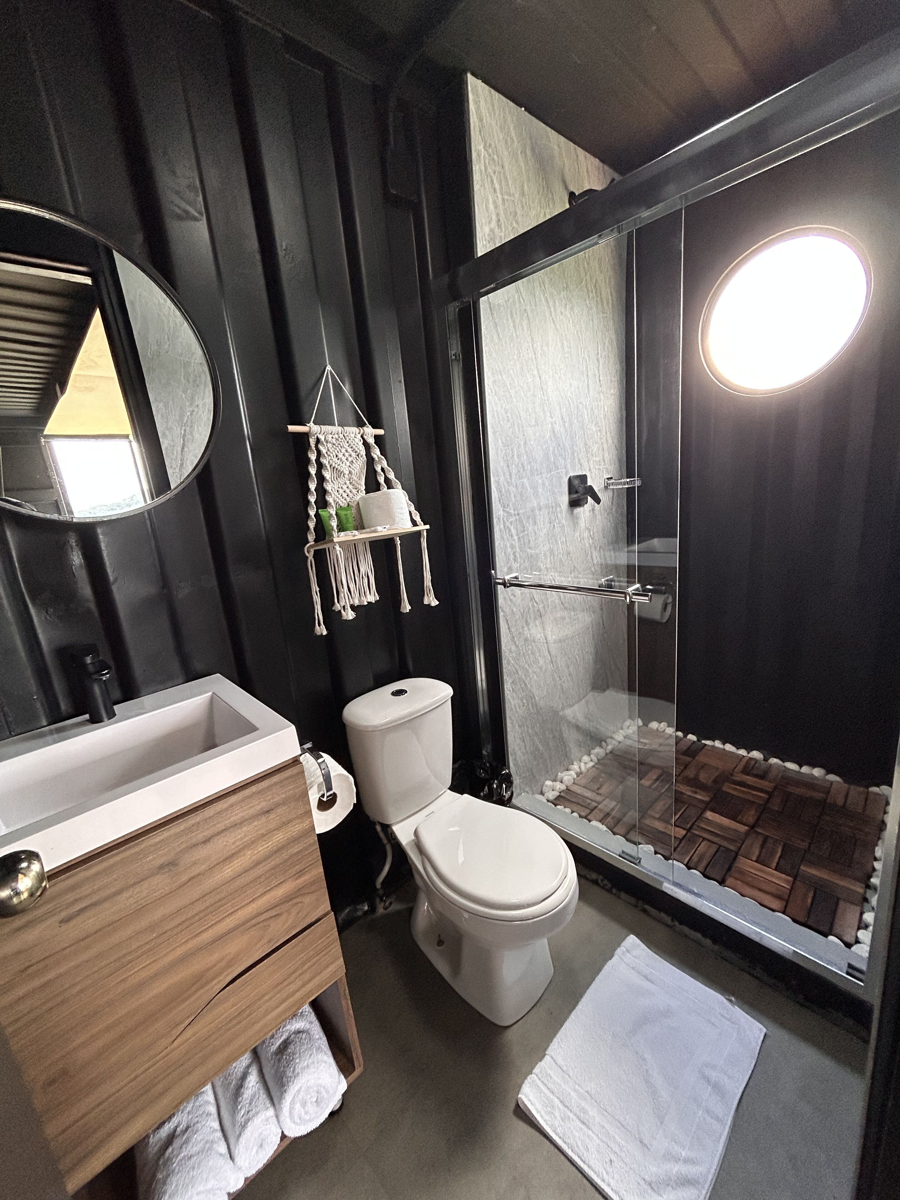 Une salle de bain avec un meuble-lavabo en bois, des toilettes, une douche avec une porte en verre et un miroir rond.