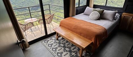 A bedroom with a large bed, a wooden bench, a small table, and a view of a mountainous landscape through large windows.