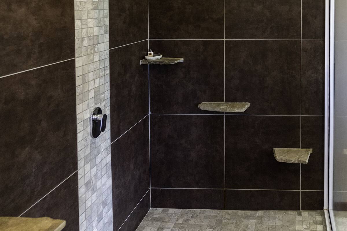 Een douche met donkere tegels, een houten plank en een betegelde vloer.