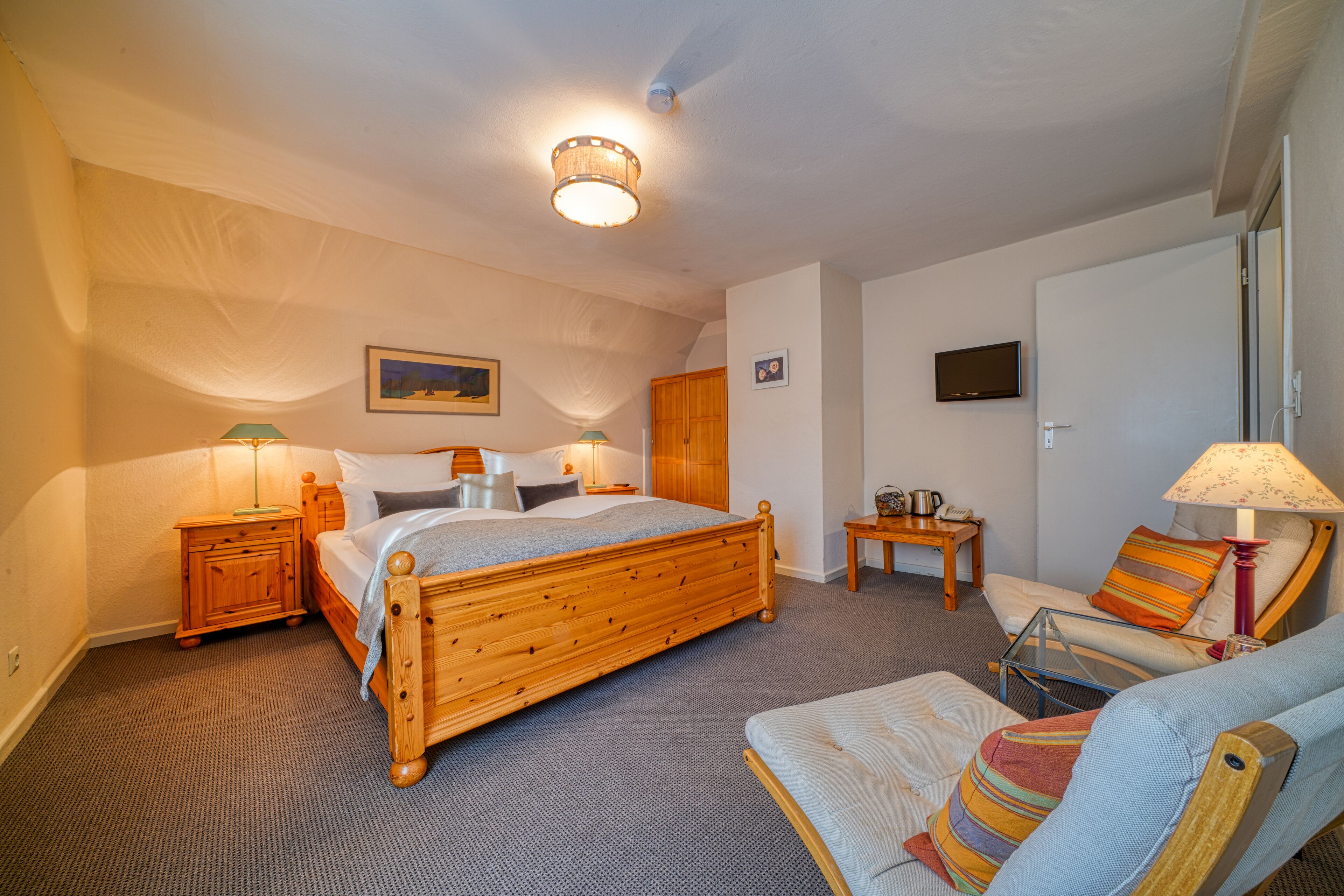 A bedroom with a wooden bed, two bedside tables with lamps, a chair, a small table, and a television on the wall.