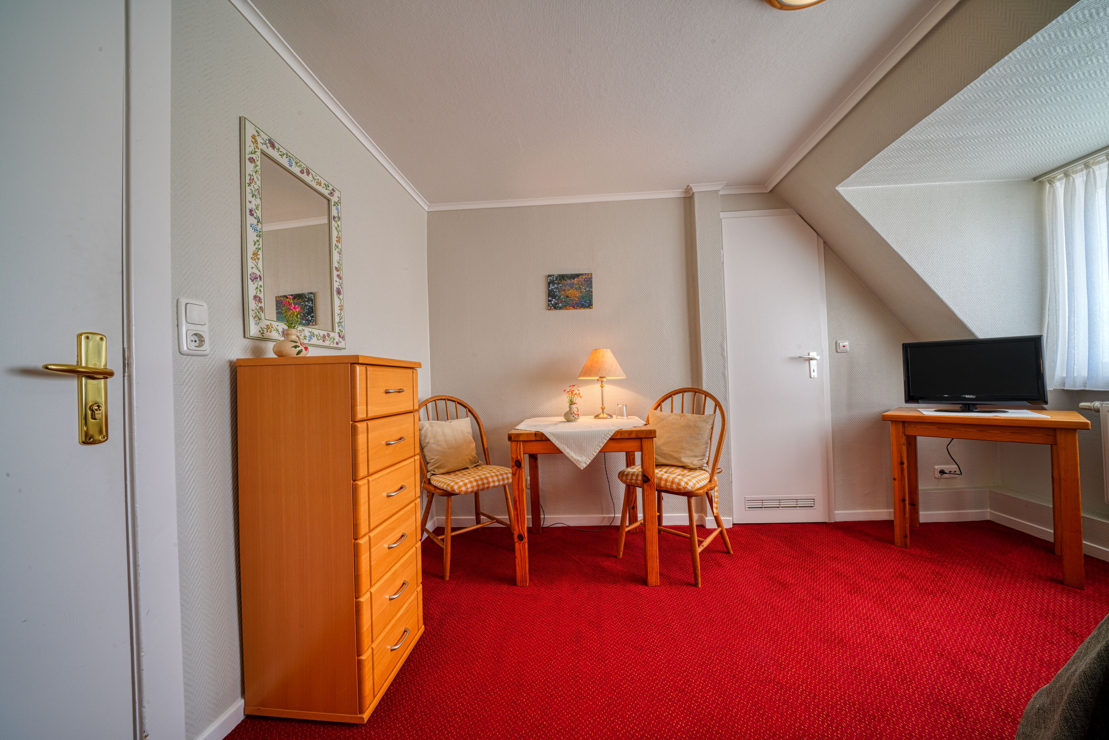 Une pièce avec une moquette rouge, une commode en bois, deux chaises, une table avec une lampe, un téléviseur et un tableau accroché au mur.