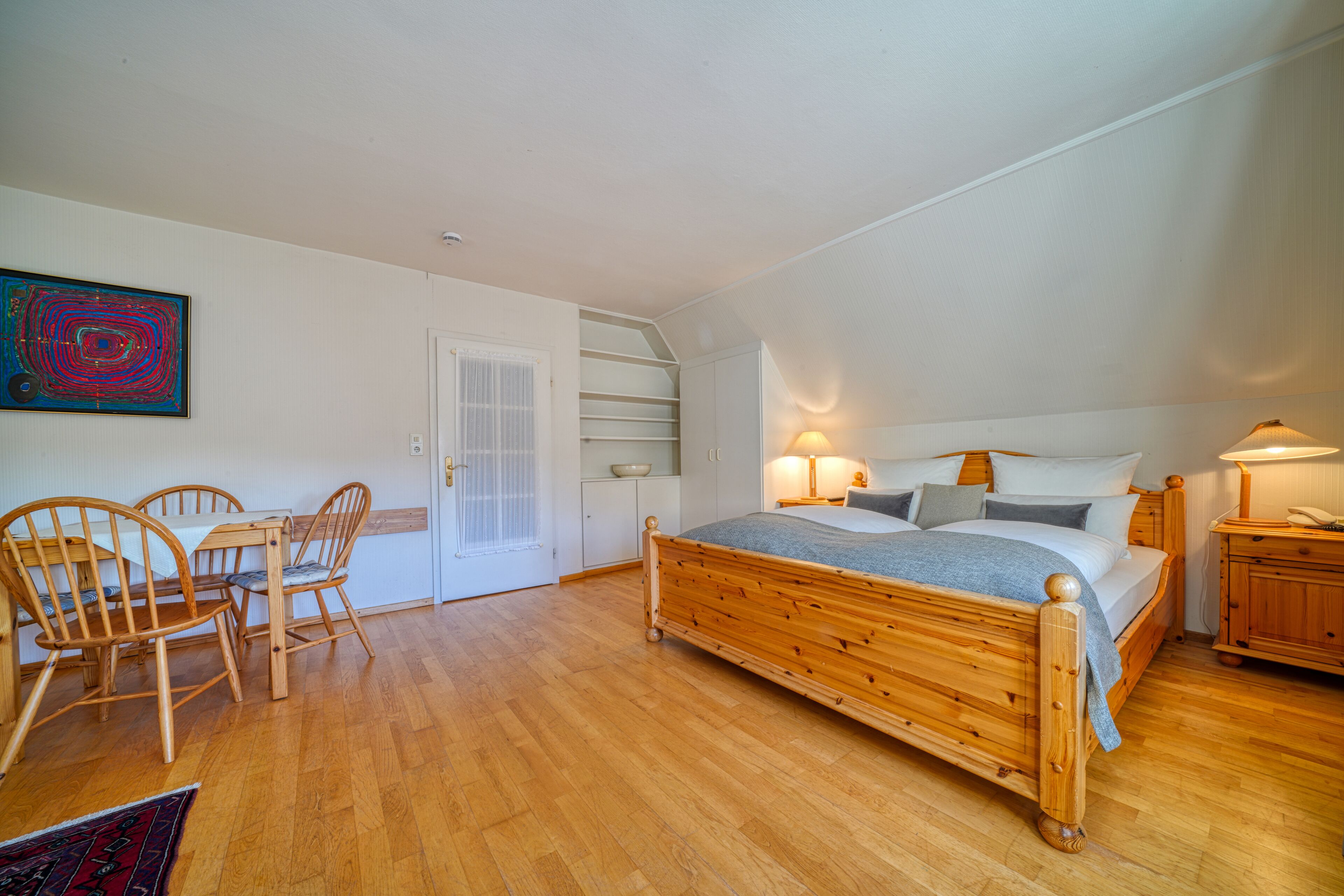 Ein Schlafzimmer mit einem hölzernen Bett, einem Esstisch mit Stühlen, einer weißen Tür und einem Gemälde an der Wand.