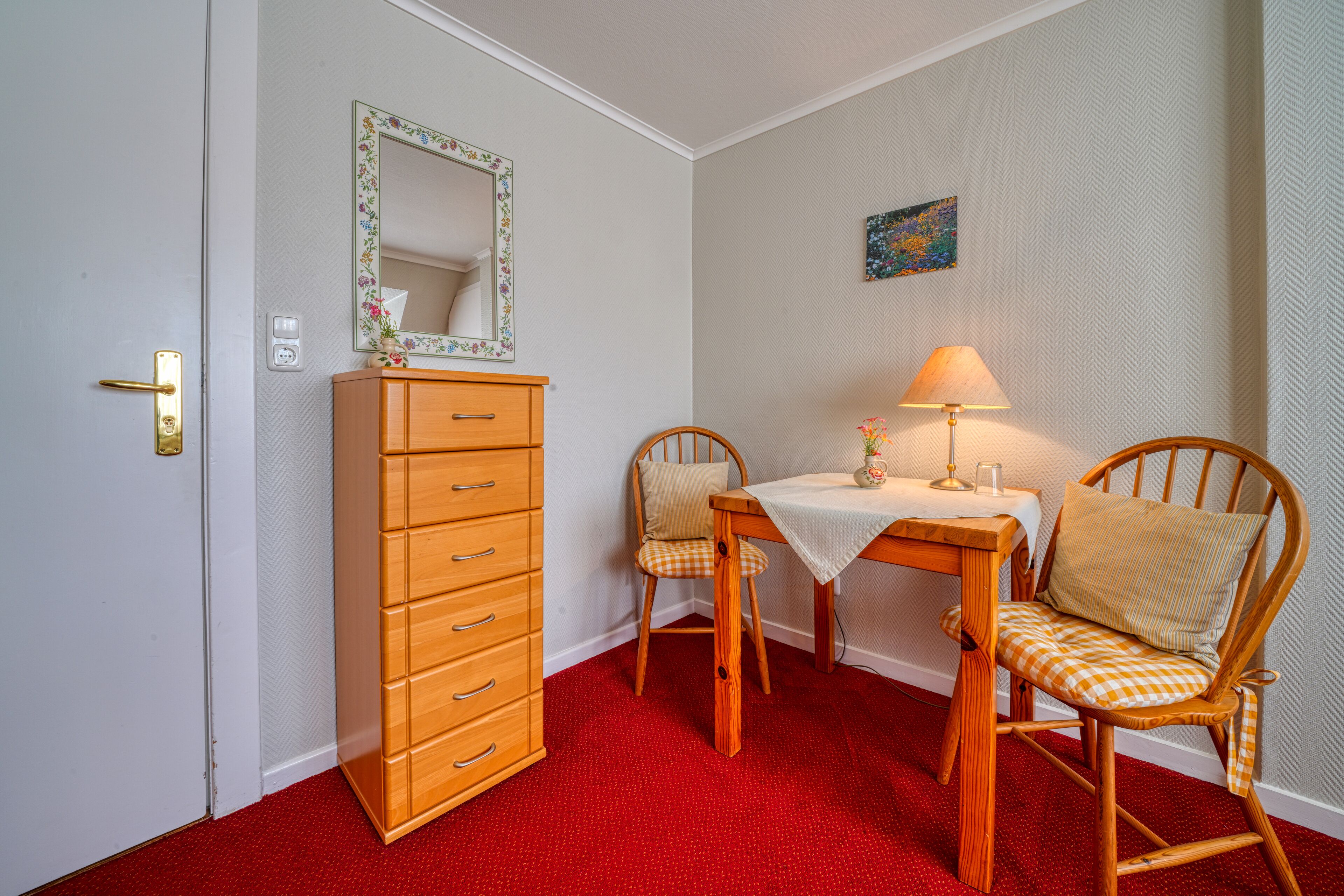 A room with a wooden dresser, two chairs, a table, a lamp, and a mirror on the wall.