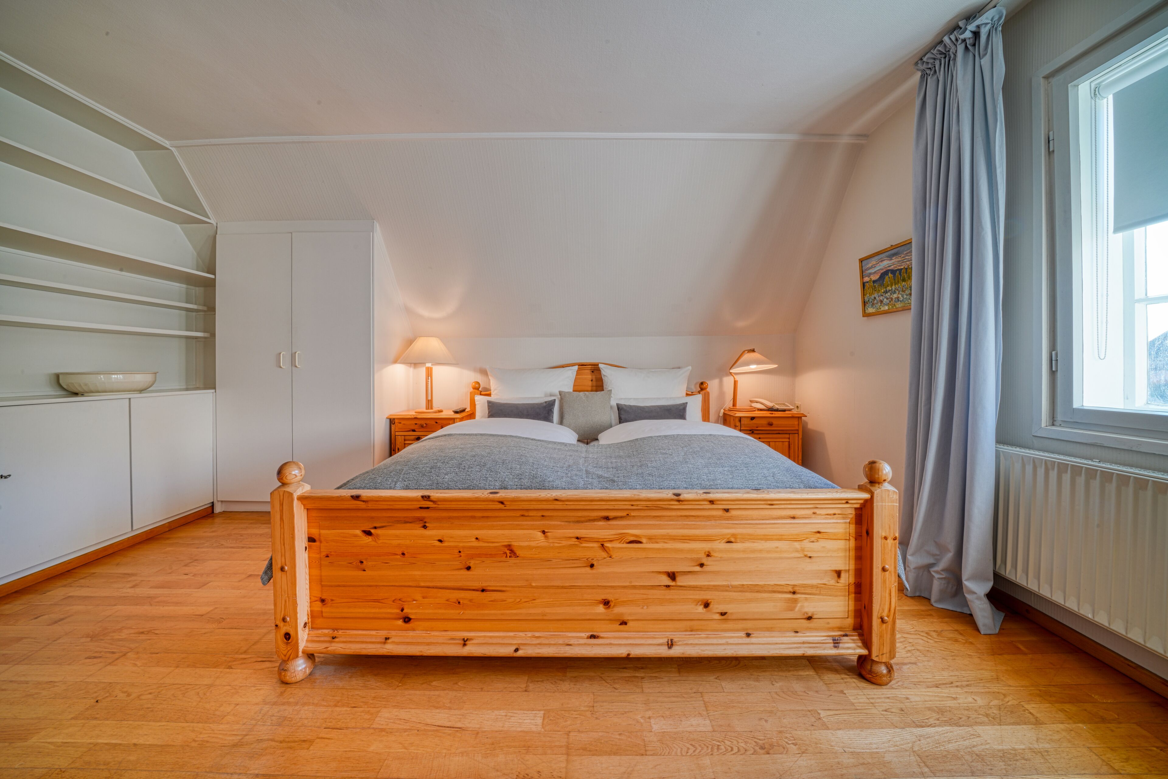 Une chambre à coucher avec un lit en bois, des murs blancs, une fenêtre avec des rideaux et une table de chevet avec des lampes.