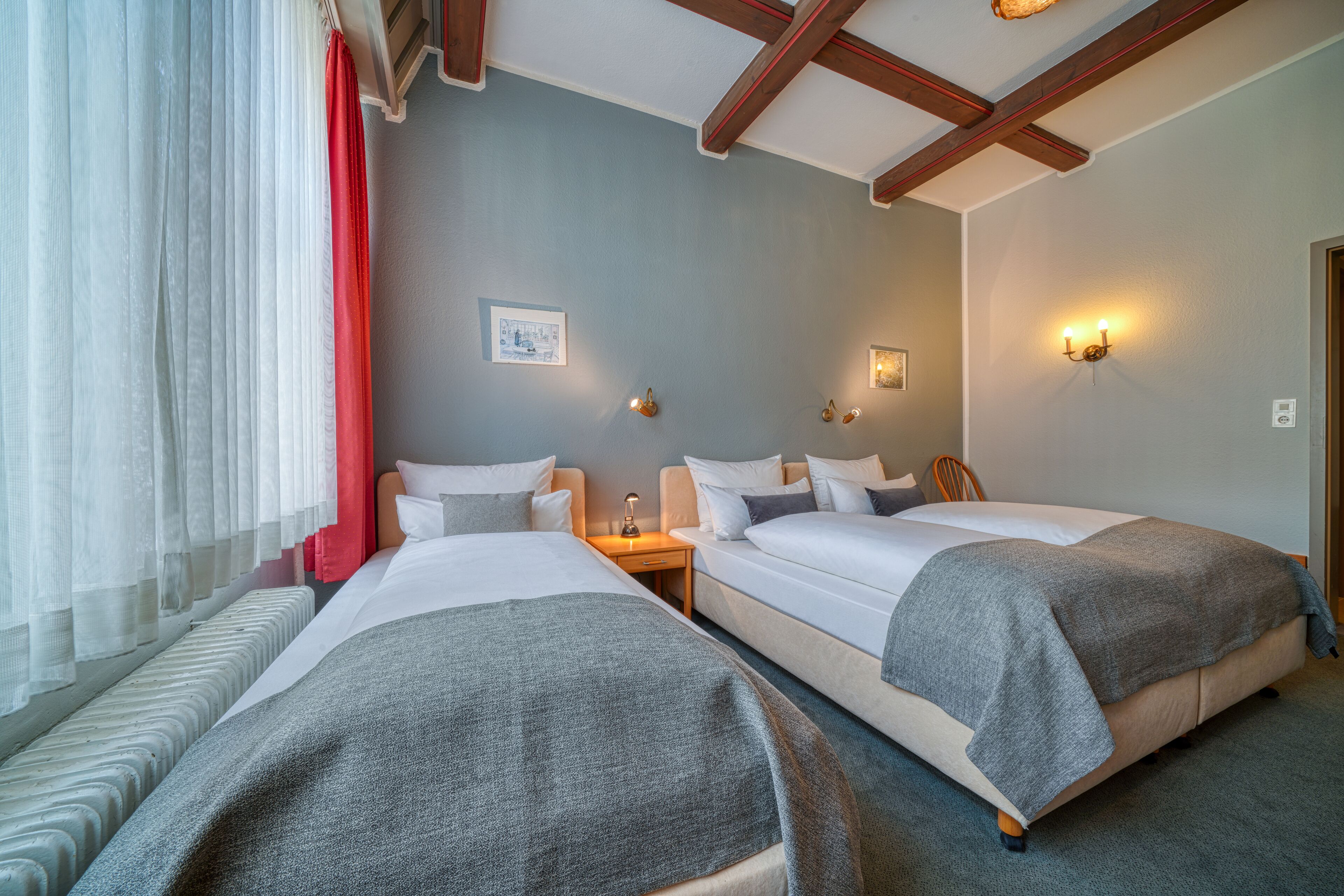 A hotel room with two beds, a window with sheer curtains, a picture on the wall, and a wooden ceiling.