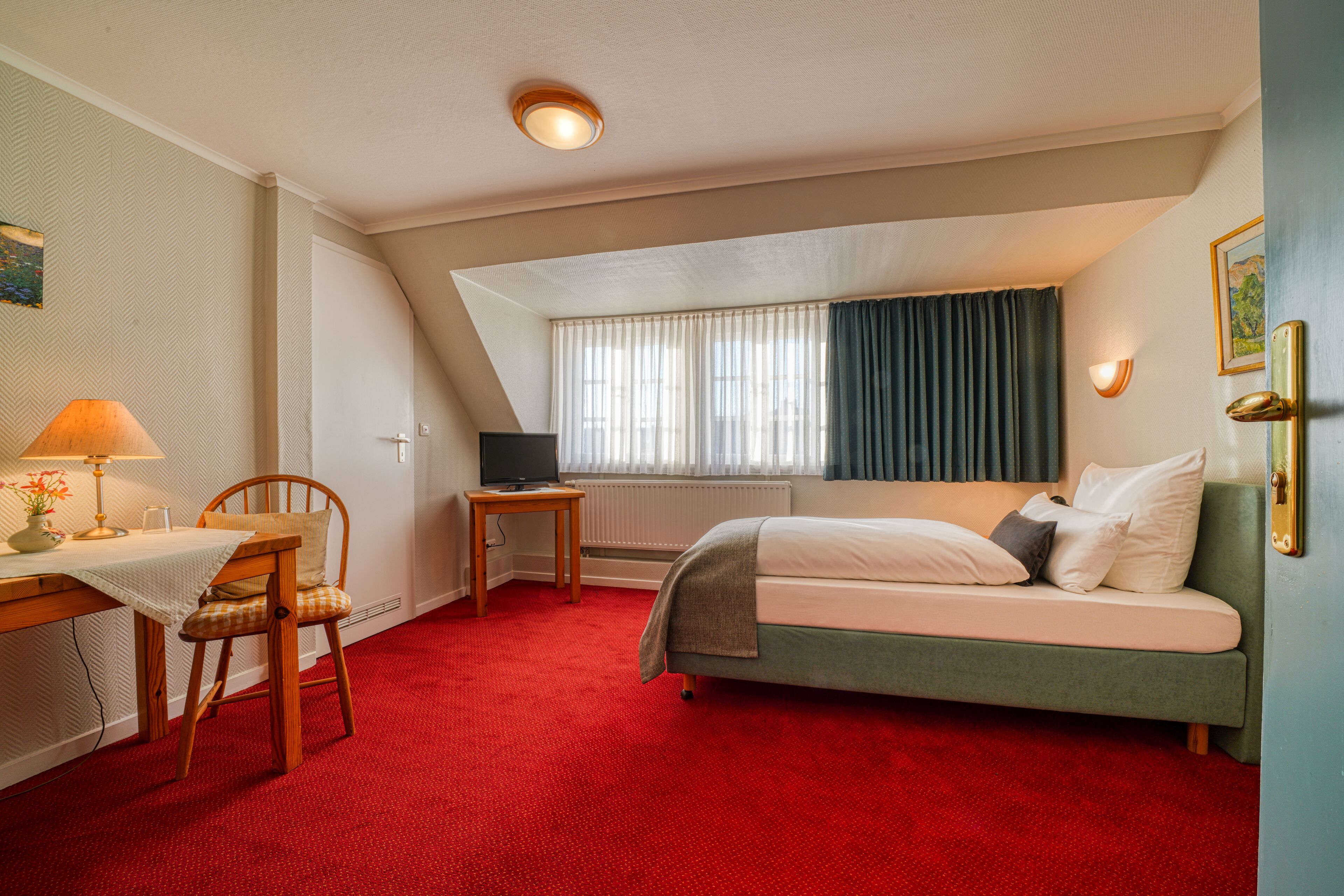 Habitación de hotel con alfombra roja, cama, escritorio con lámpara, silla, televisión y ventana con cortinas.