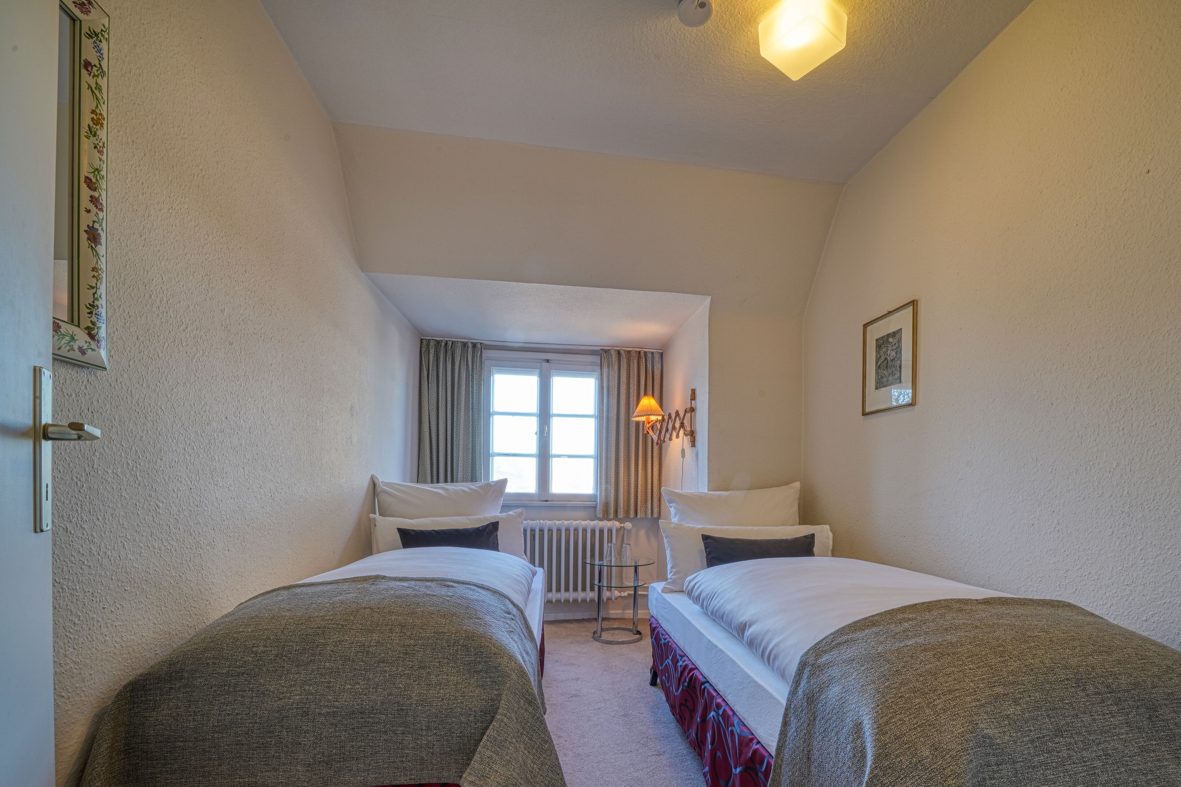 A hotel room with two single beds, a small table, a chair, a radiator, and a window with curtains.