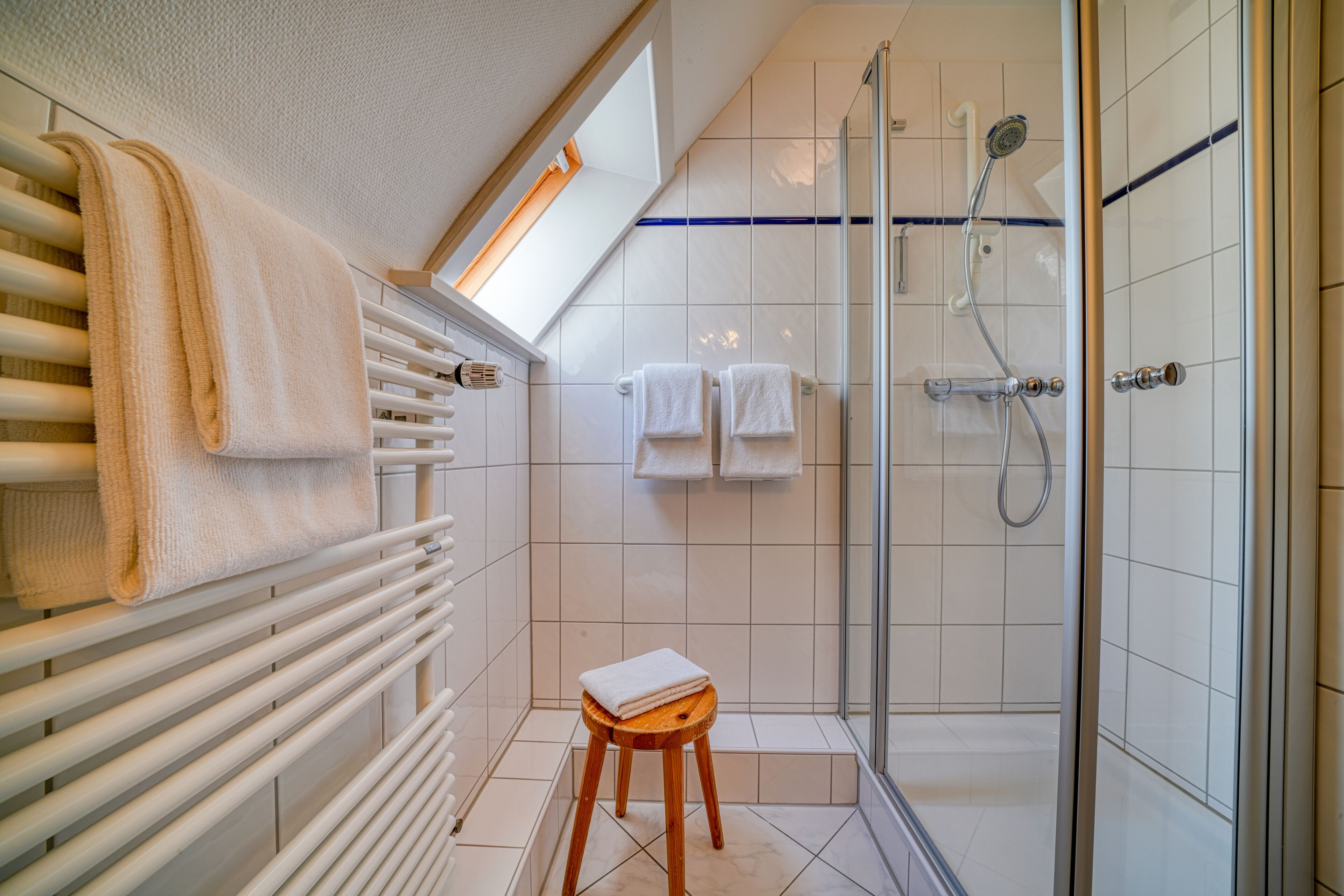 A modern bathroom with a glass shower, white tiled walls, a wooden stool, and a towel rack.
