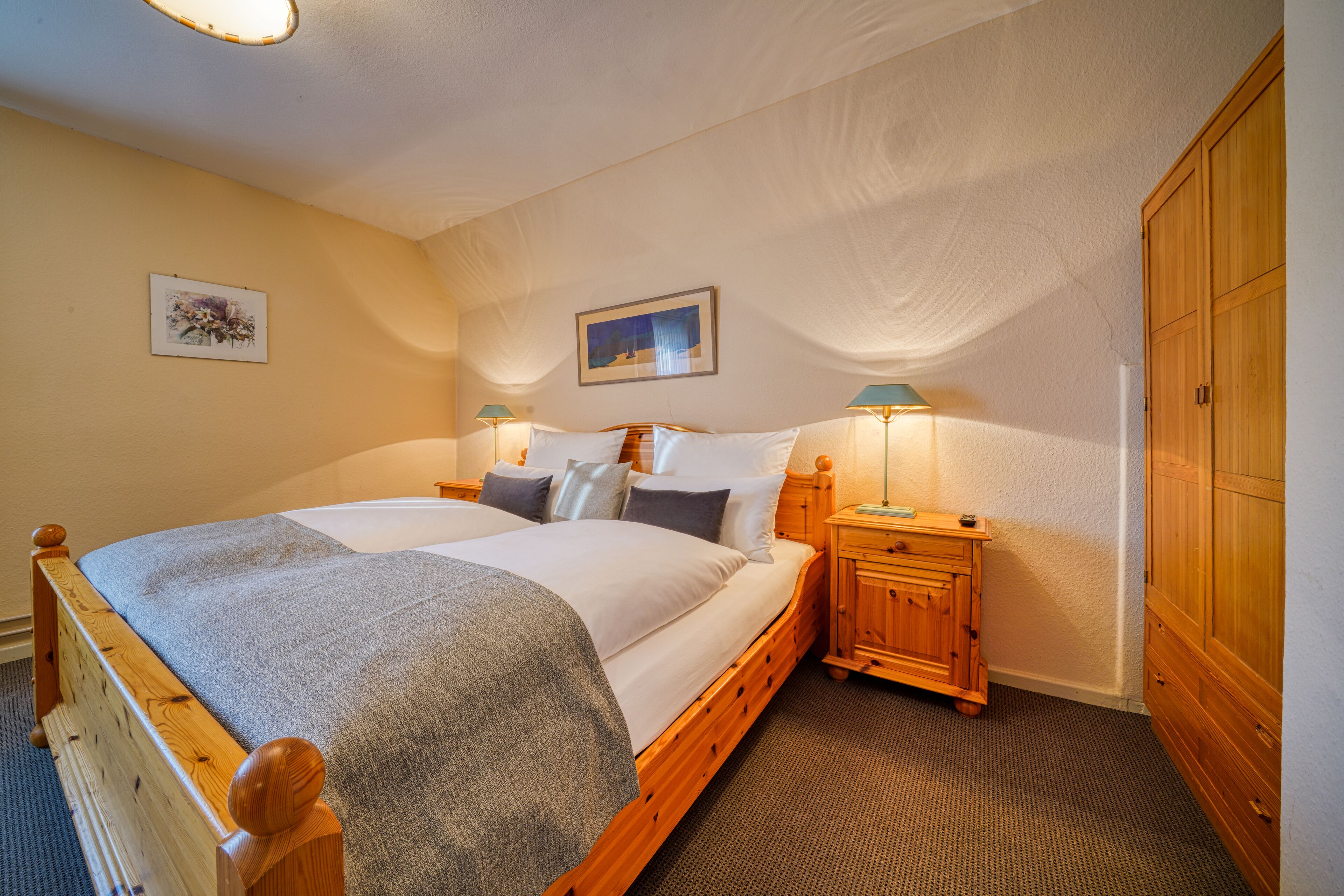 Une chambre à coucher avec un lit en bois, deux tables de chevet avec des lampes, une armoire et deux tableaux encadrés accrochés au mur.