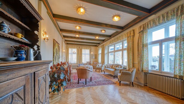Una habitación amplia con piso de madera, un ventanal con cortinas florales y una chimenea tradicional.