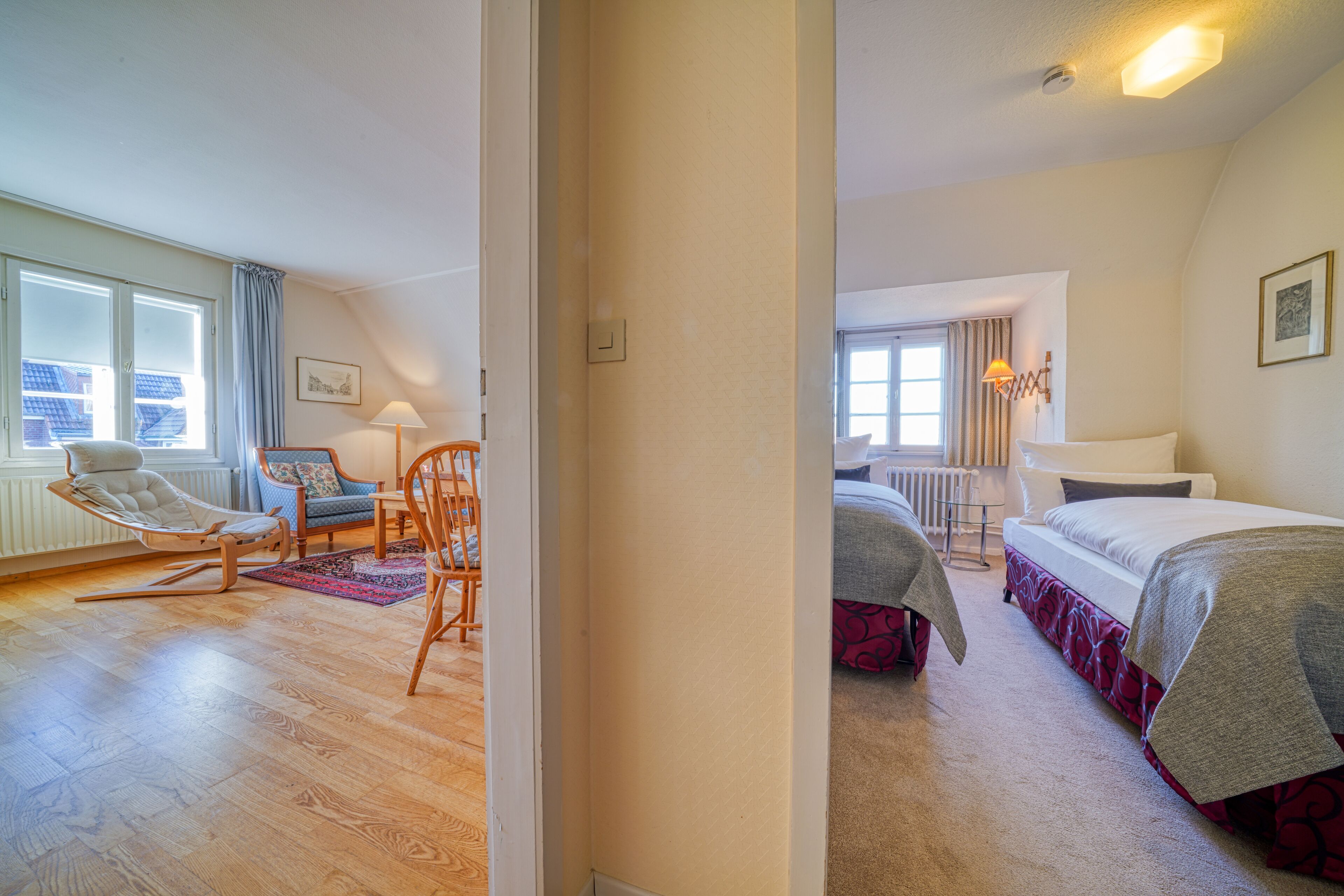 Habitación de hotel con piso de madera, una mecedora, una cama con colcha roja y blanca y una ventana con cortinas.