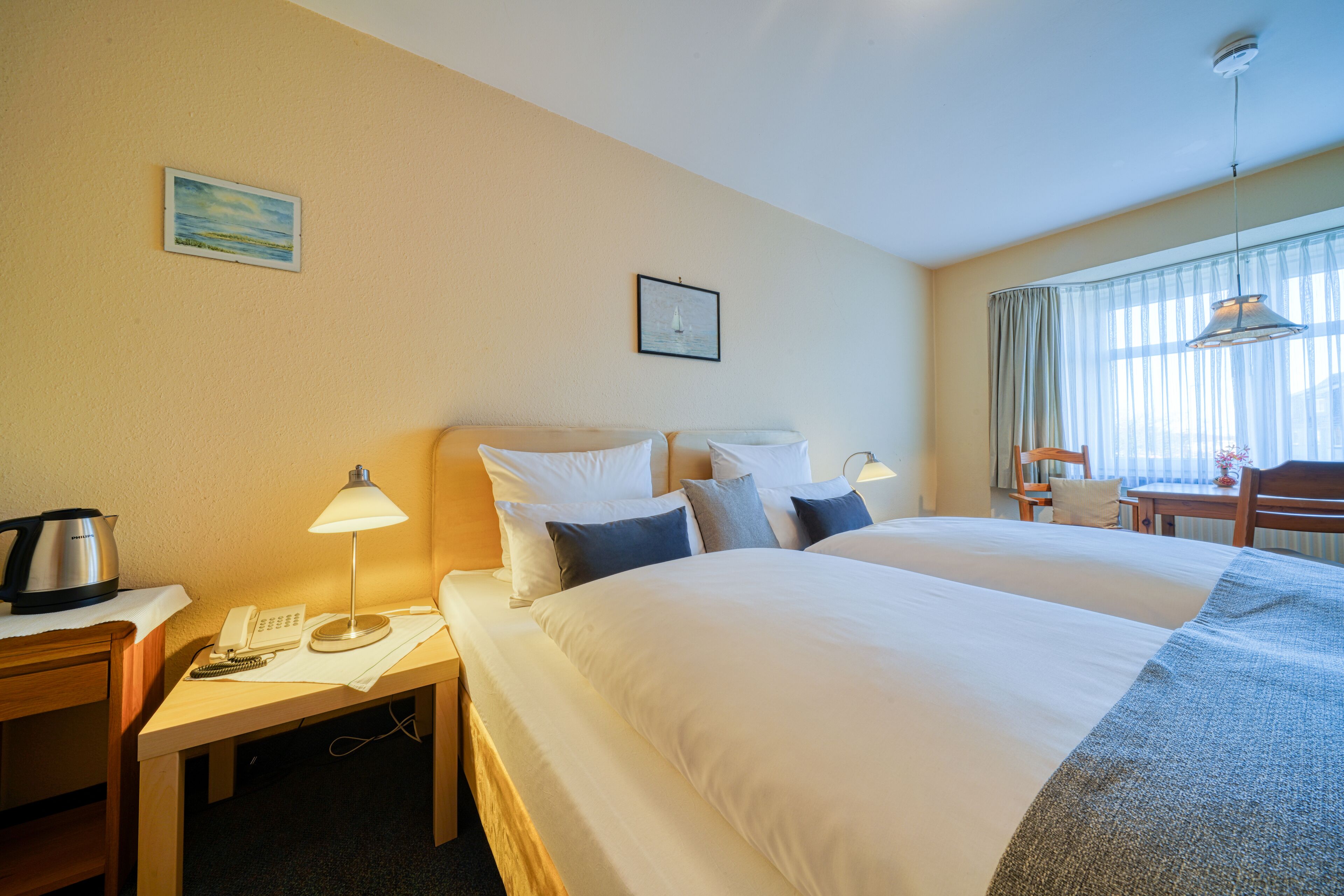 Ett hotellrum med två sängar, ett skrivbord med en telefon och en lampa, en stol och ett bord med en lampa.
