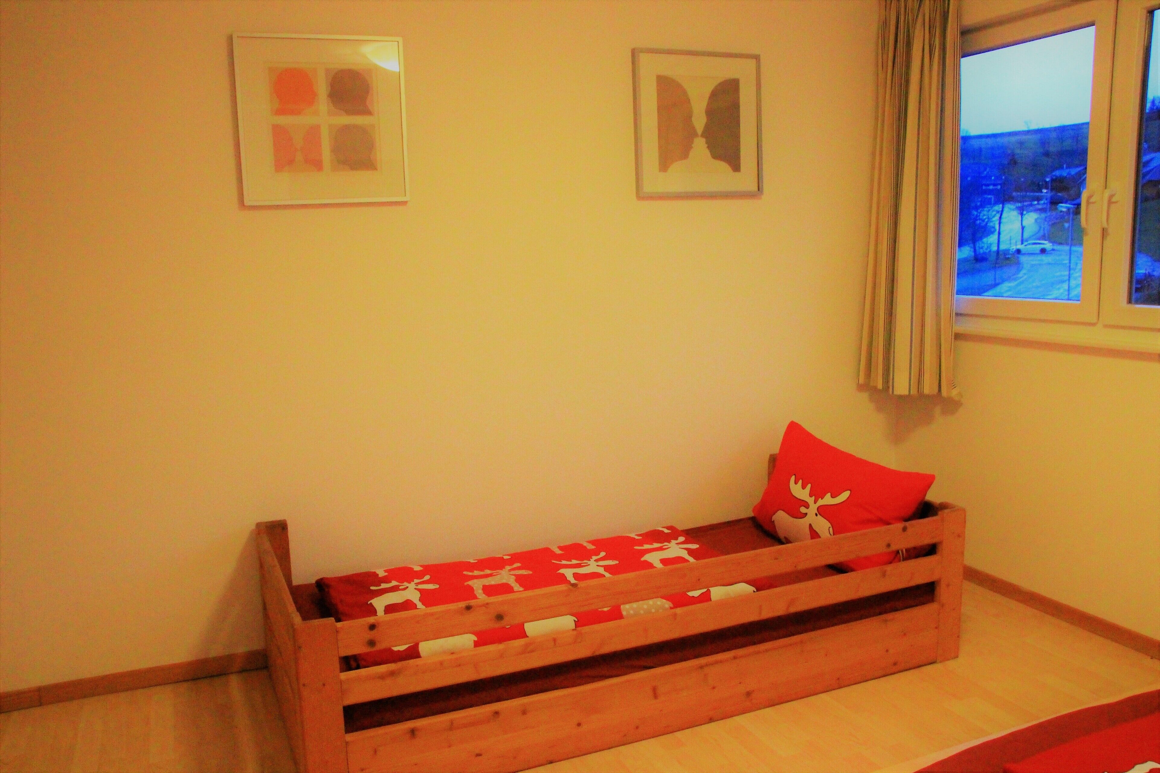 A small wooden bed with a red and white patterned bedspread and a single red pillow. There are two framed pictures on the wall above the bed.