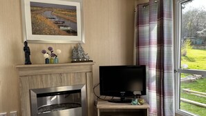 A living room with a fireplace, a flat-screen TV on a wooden stand, a framed painting above the fireplace, and a view of a green landscape through a window with curtains. - Vacation chalet on a campsite (Windeck)