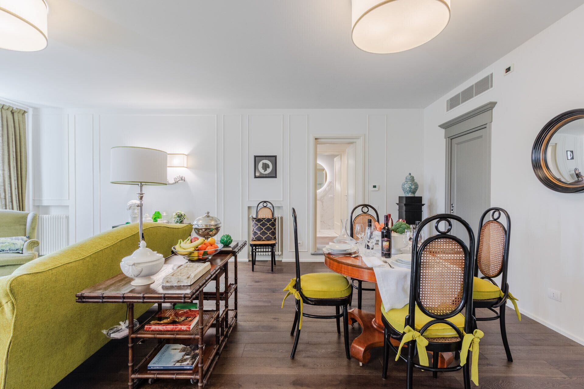 Un coin repas avec une table dressée pour quatre personnes, entourée de chaises dotées de coussins jaunes. Un canapé vert est placé au premier plan, et une lampe est visible sur une table d’appoint.