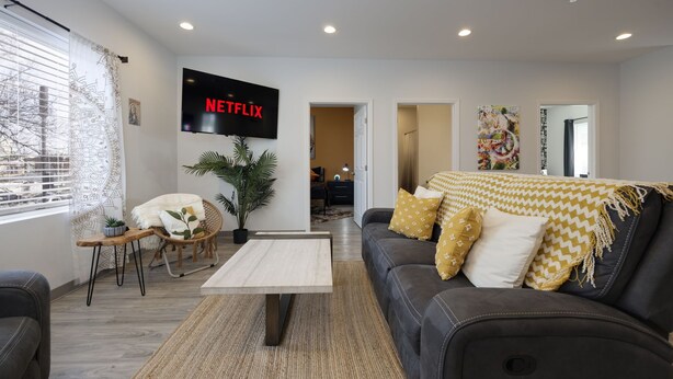 A living room with a black leather sofa, a coffee table, a TV mounted on the wall, and a potted plant. - TWO Condos! Side by Side Curated Suites! Sleeps 18! Parking! (Nashville)