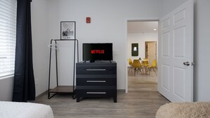 A modern hotel room with a black TV on a stand, a wardrobe, a bed, and a view into the dining area. - TWO Condos! Side by Side Curated Suites! Sleeps 18! Parking! (Nashville)