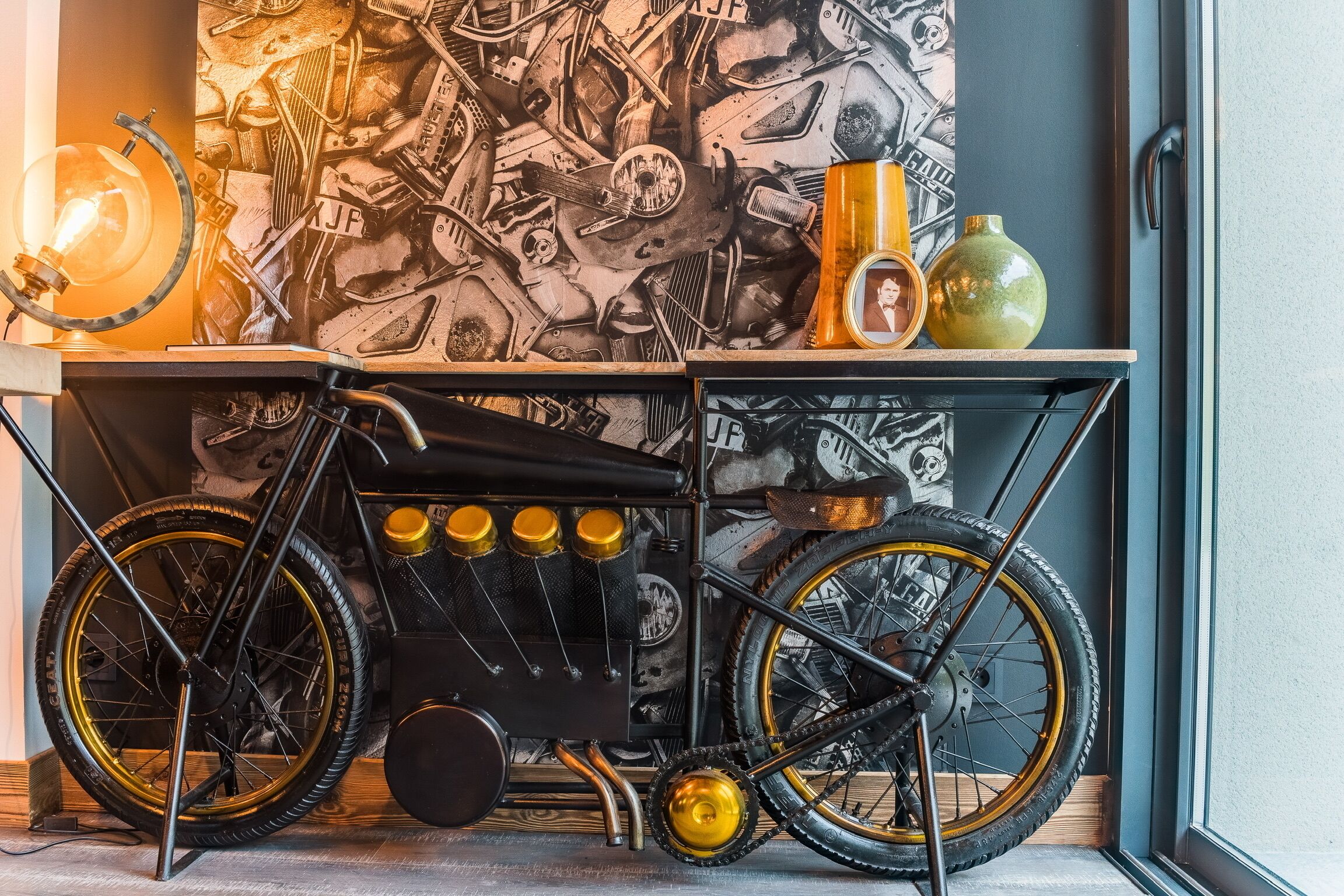 A bicycle cart with a vintage design, a light bulb, and a vase with a picture on a table against a wall with a mechanical pattern.