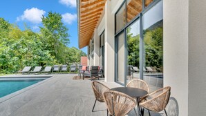 Un espace piscine extérieur moderne, doté de chaises longues et d’une table, entouré de murs de verre et de poutres en bois au plafond.