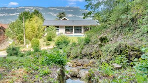 Une maison moderne dotée d’une grande baie vitrée, entourée d’une végétation luxuriante et avec une chaîne de montagnes en toile de fond.