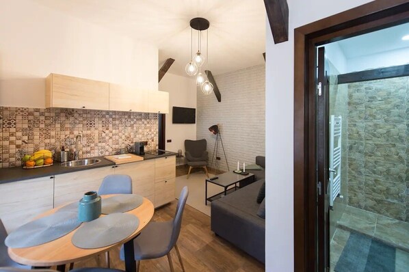 A compact kitchen with a tiled backsplash, a dining area with a table and chairs, a gray sofa, and a stone wall adjacent to a sliding glass door leading to a stone patio.