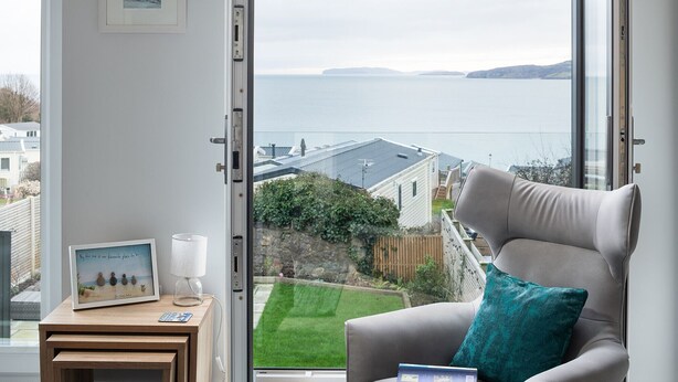 Una habitación con vista al mar, una silla con cojín, una mesita con una lámpara y un cuadro enmarcado, y un libro sobre la silla.