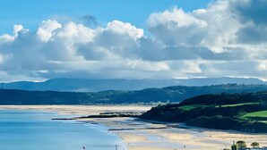 Un paisaje costero con playa, acantilados y una cadena montañosa distante bajo un cielo parcialmente nublado.