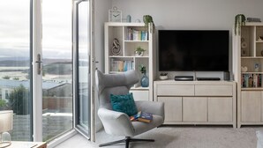 Una sala de estar moderna con un sillón gris, una estantería, un televisor de pantalla plana y vistas a través de un ventanal.