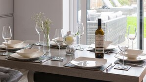 Una mesa de comedor dispuesta con platos blancos, copas de vino y una botella de vino, con vistas a un jardín y un área de asientos al aire libre.