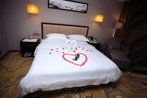 Ein Hotelzimmer mit einem ordentlich bezogenem Bett, einer herzförmigen Dekoration mit Rosenblättern und einem Bild an der Wand.