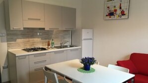 Une cuisine compacte avec un coin repas, des meubles blancs, un petit réfrigérateur et un canapé rouge.