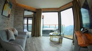 Un salón moderno con sofá, una mesa de centro y televisor. Amplias ventanas ofrecen vistas al exterior.