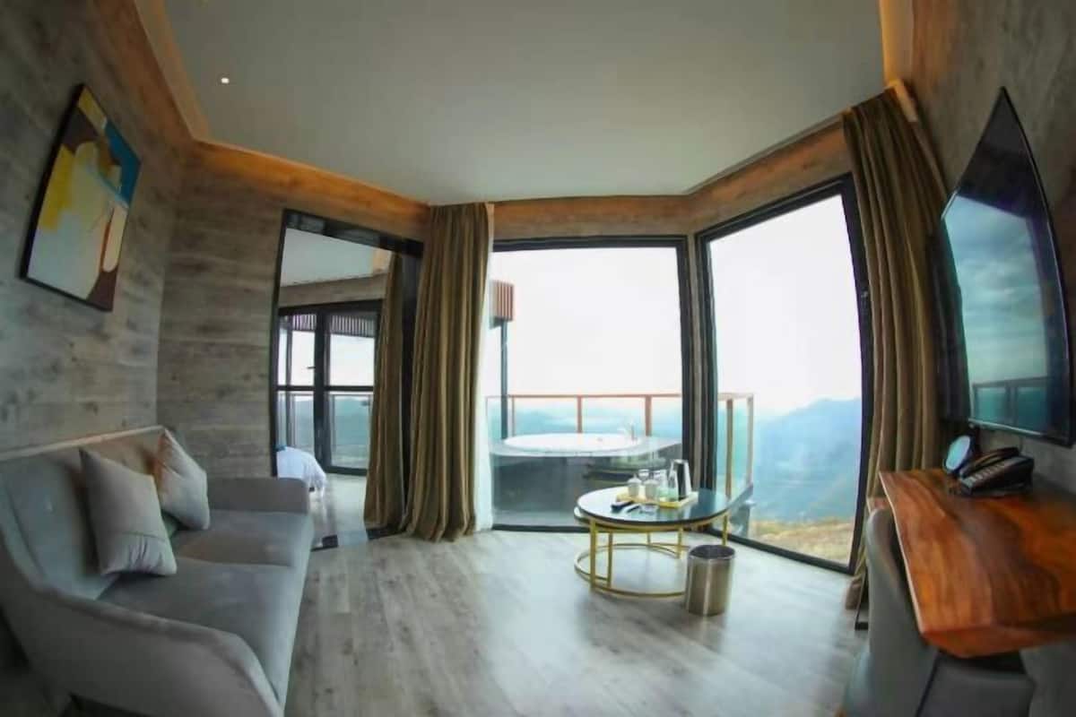 A modern living room with a sofa, a coffee table, and a TV. There are large windows offering a view of the outdoors.