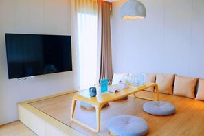 Ein modernes Wohnzimmer mit einem wandmontierten Flachbildfernseher, einem Couchtisch aus Holz mit Glasplatte, zwei grauen Hocker und einem großen Fenster mit Vorhängen.