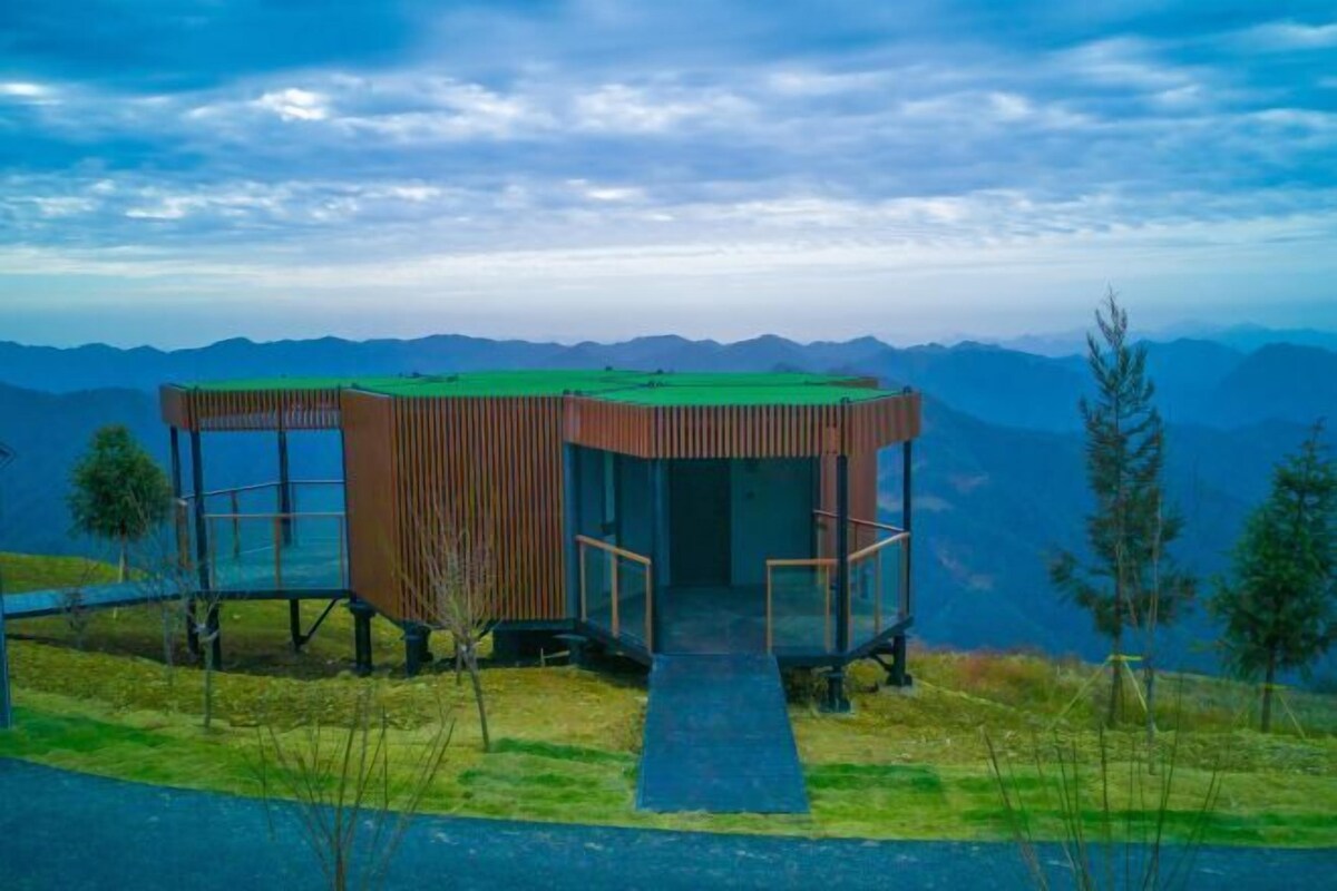 Eine moderne, hölzerne Hütte mit Glaseinfriedung, gelegen auf einem grasbewachsen Hügel mit Bergblick.