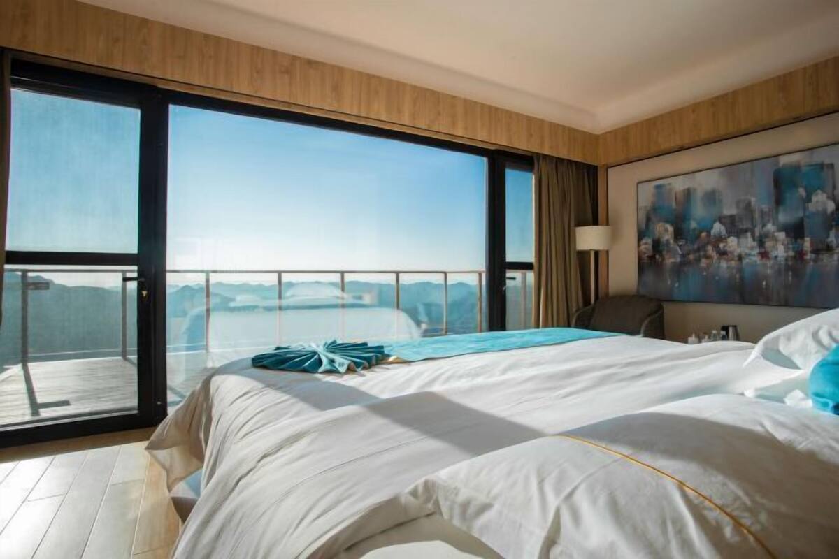 A modern hotel room with a large bed, a painting on the wall, and a balcony with a view of the mountains.