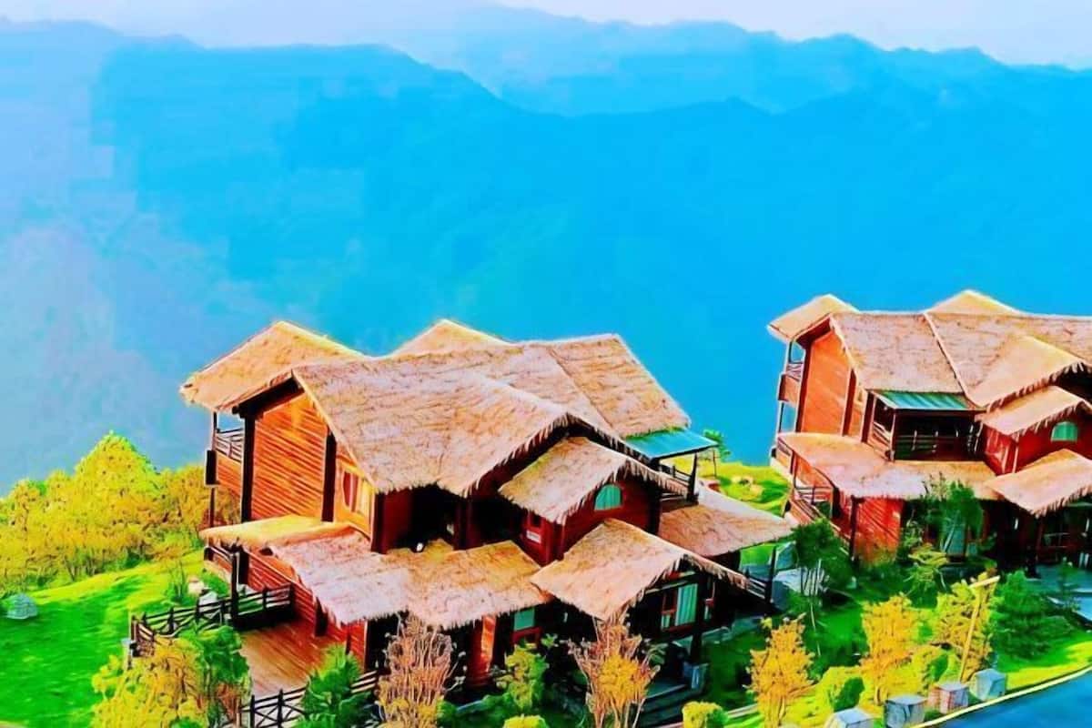 A cluster of rustic wooden cabins with thatched roofs, surrounded by lush greenery and overlooking a mountainous landscape.