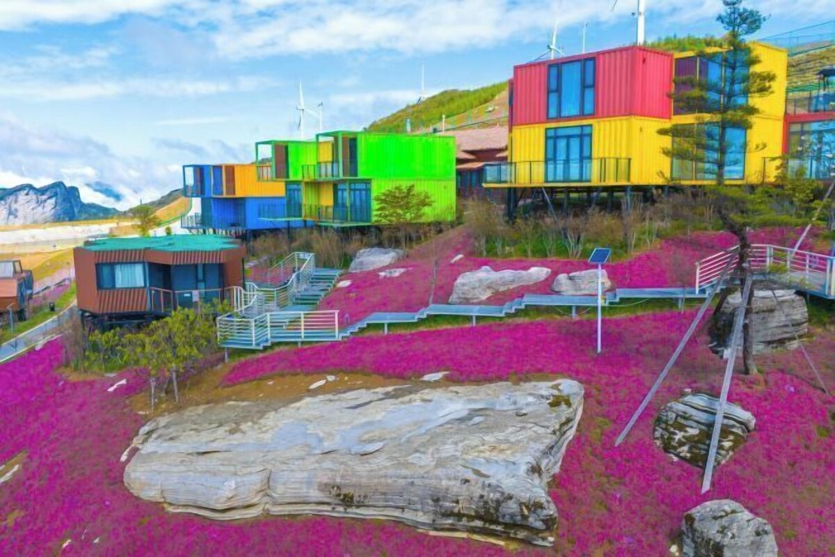 Ein farbenfrohes, modernes Gebäude mit mehreren Ebenen und Balkonen, gelegen an einem Hang mit leuchtend rosa Blumen und einem großen Felsen im Vordergrund.