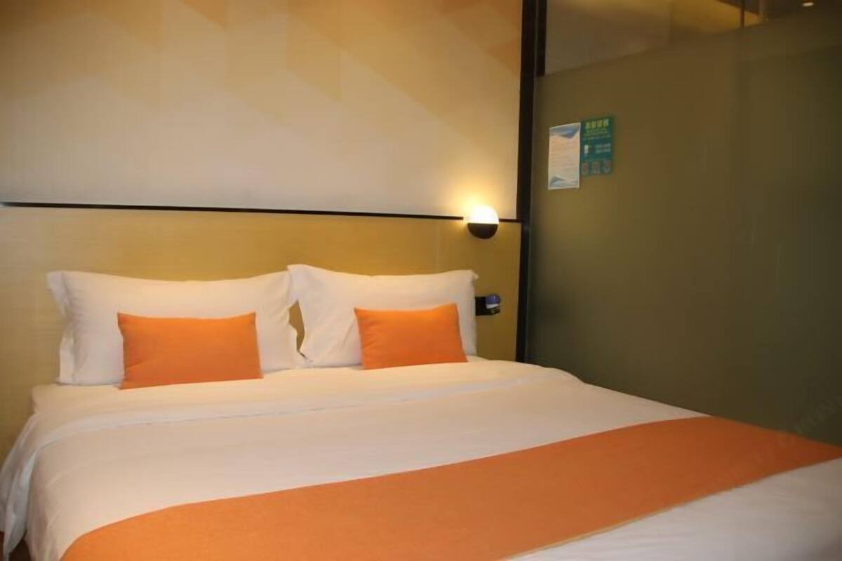 Une chambre d’hôtel avec un lit, une literie blanche et orange, une lampe fixée au mur et un panneau vert sur le mur.