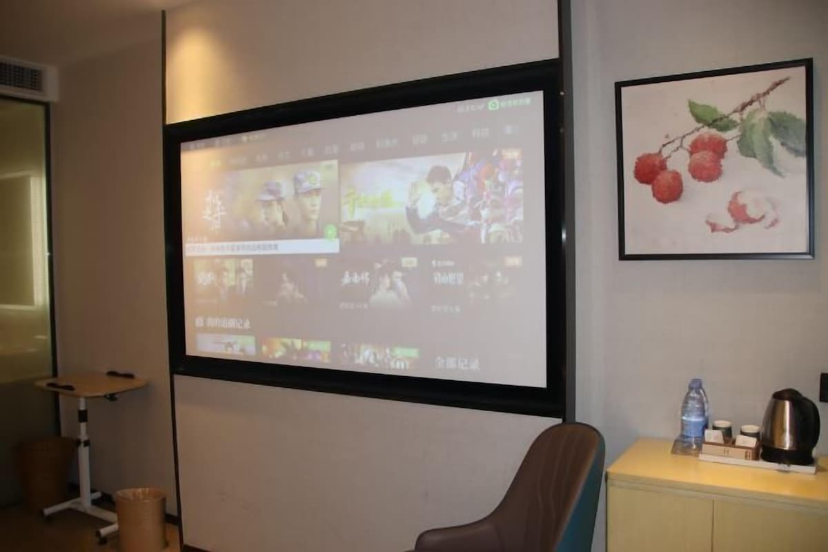 A room with a projection screen displaying a video, a framed painting of fruit, a desk with a water bottle, and a coffee maker.