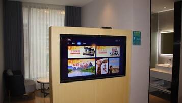 A hotel room with a large digital display on the wall, a small round table, a chair, and a window with curtains.