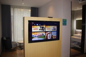 A hotel room with a large digital display on the wall, a small round table, a chair, and a window with curtains.