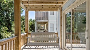A wooden deck with a swing, a glass door, and a view of a building with a balcony. - Corolla Premier: Short Walk to Beach | EV Charger (Corolla)