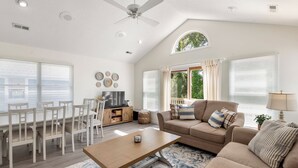 A spacious living room with a high ceiling, a ceiling fan, a dining area with a wooden table and chairs, a beige sofa with cushions, and a TV on a wooden cabinet. - Corolla Premier: Short Walk to Beach | EV Charger (Corolla)