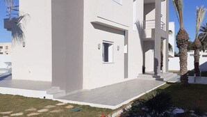 A modern white building with a flat roof, a covered entrance, and a small garden area with palm trees and shrubs. - Villa Sonia with infinity pool (djerba)